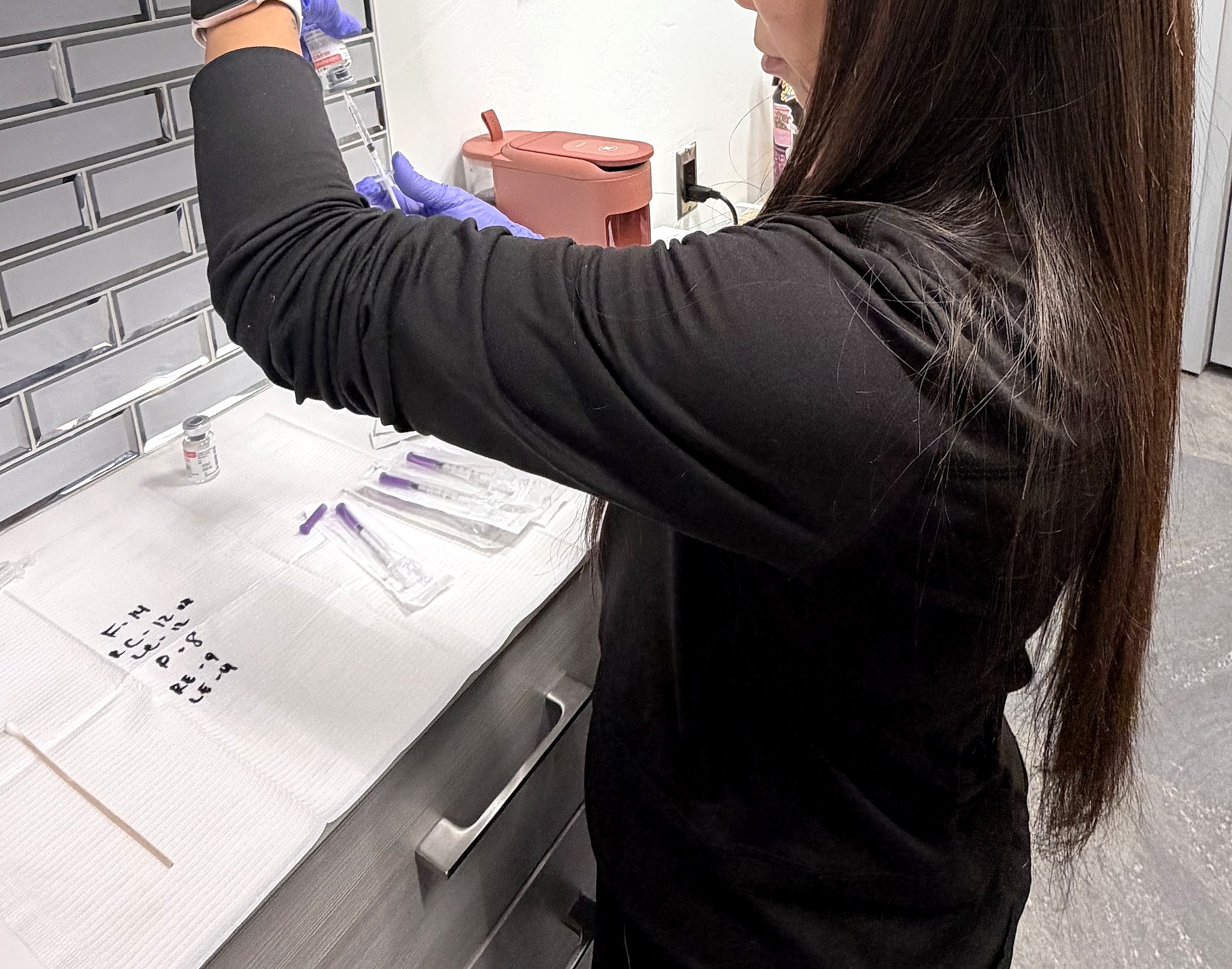 Kristine in gloves drawing up a treatment, syringes laid out on the counter